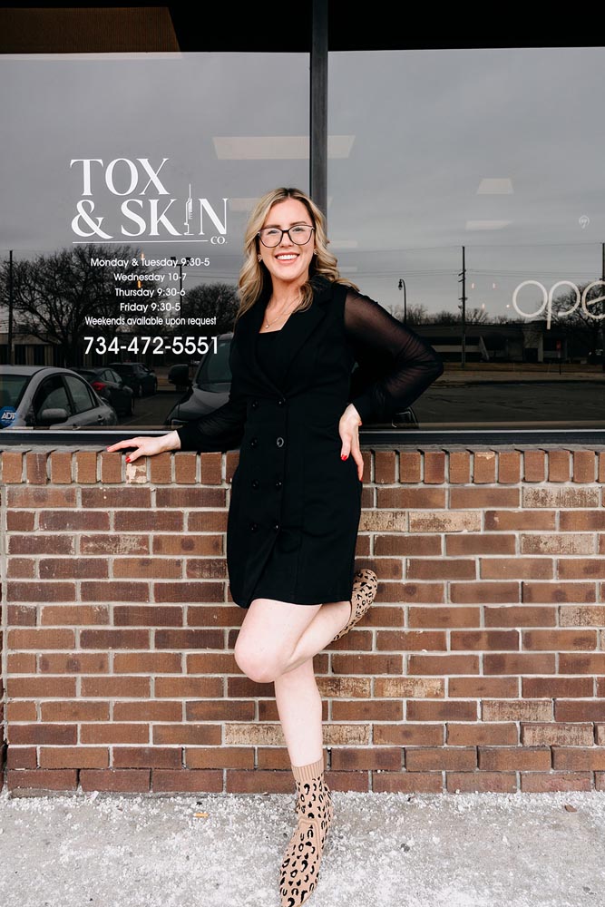 Woman in black dress smiles outside Tox & Skin Co., leaning against a brick wall.