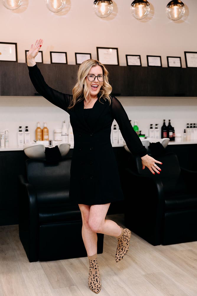A person with glasses and a black dress striking a playful pose in a salon.