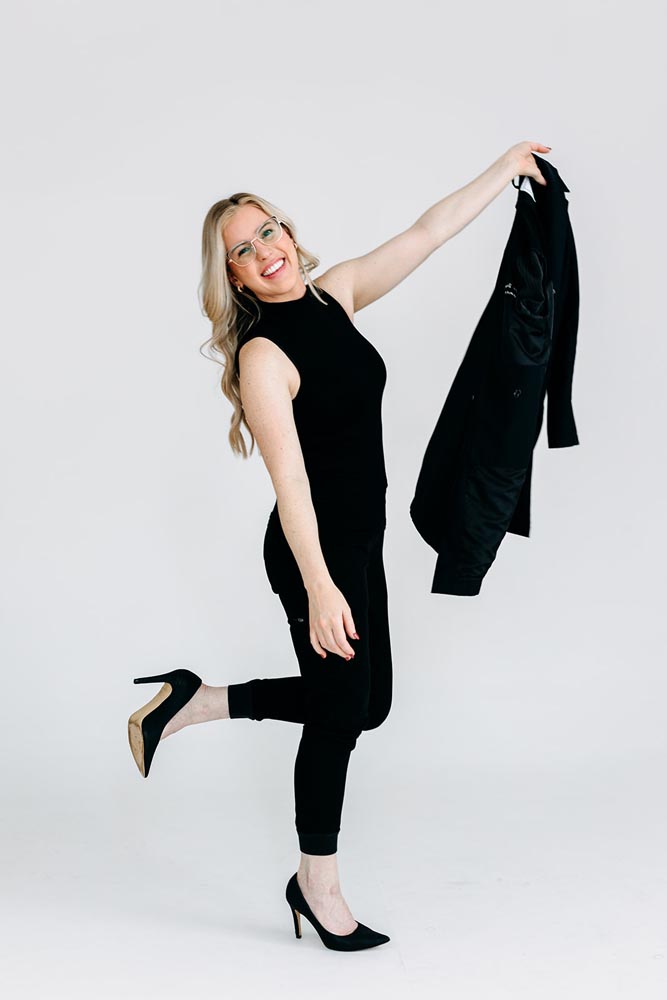 A woman in black attire smiling and holding a jacket, posing on one foot.