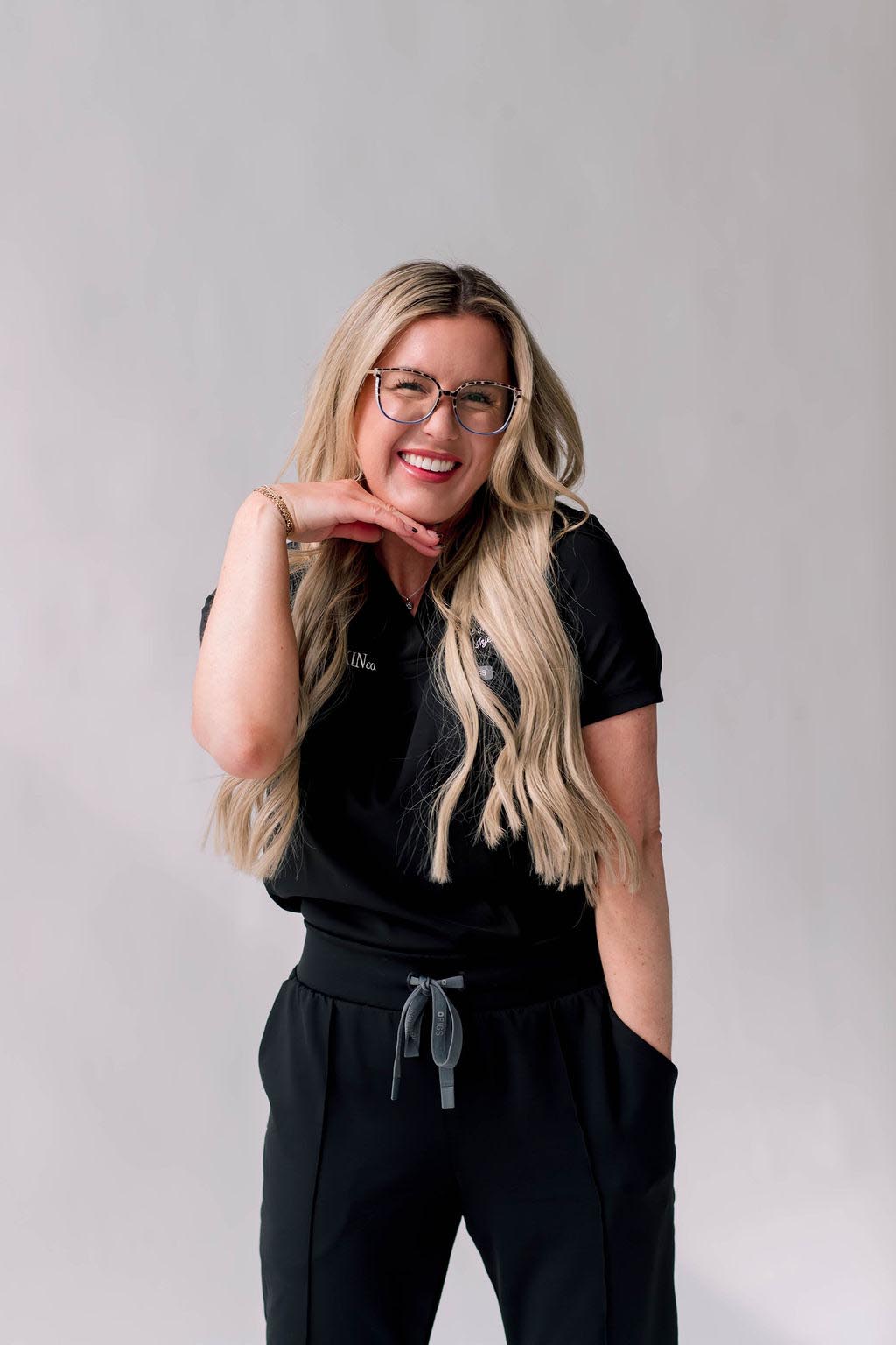 A woman with long blonde hair smiling and posing with a hand under her chin in a black outfit.
