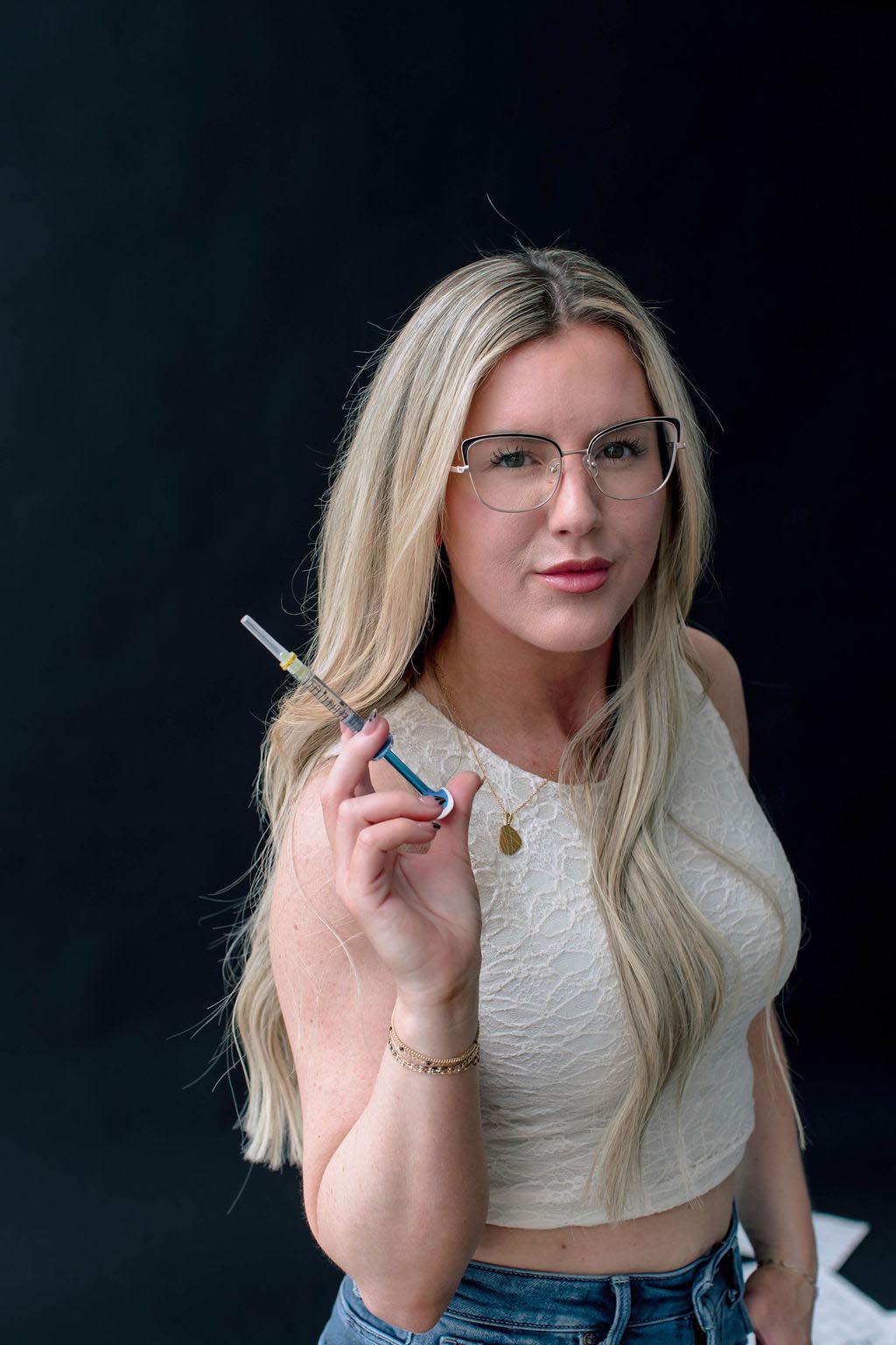 A woman with glasses holding a syringe against a dark background.