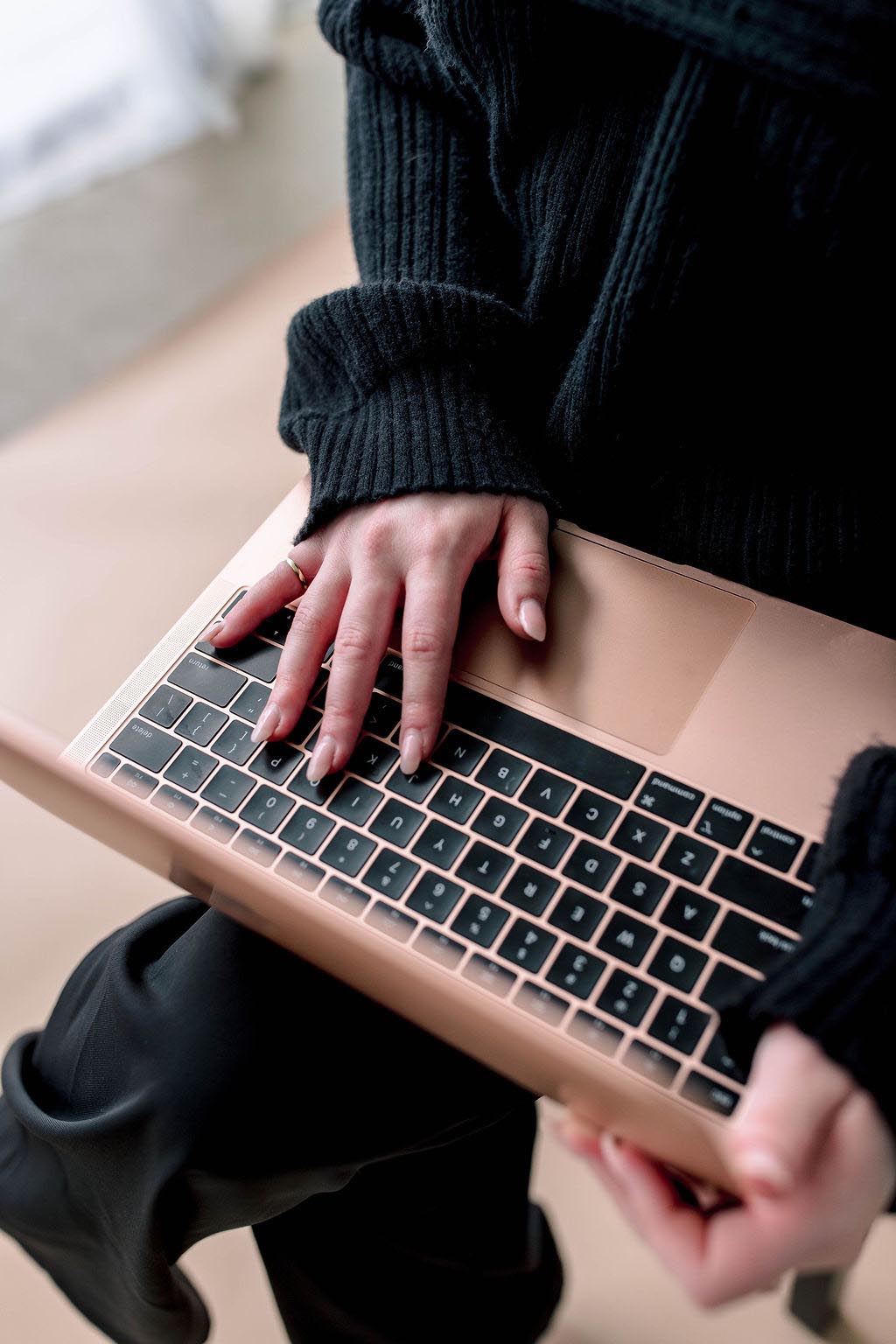 A person typing on a rose gold laptop wearing a black sweater.