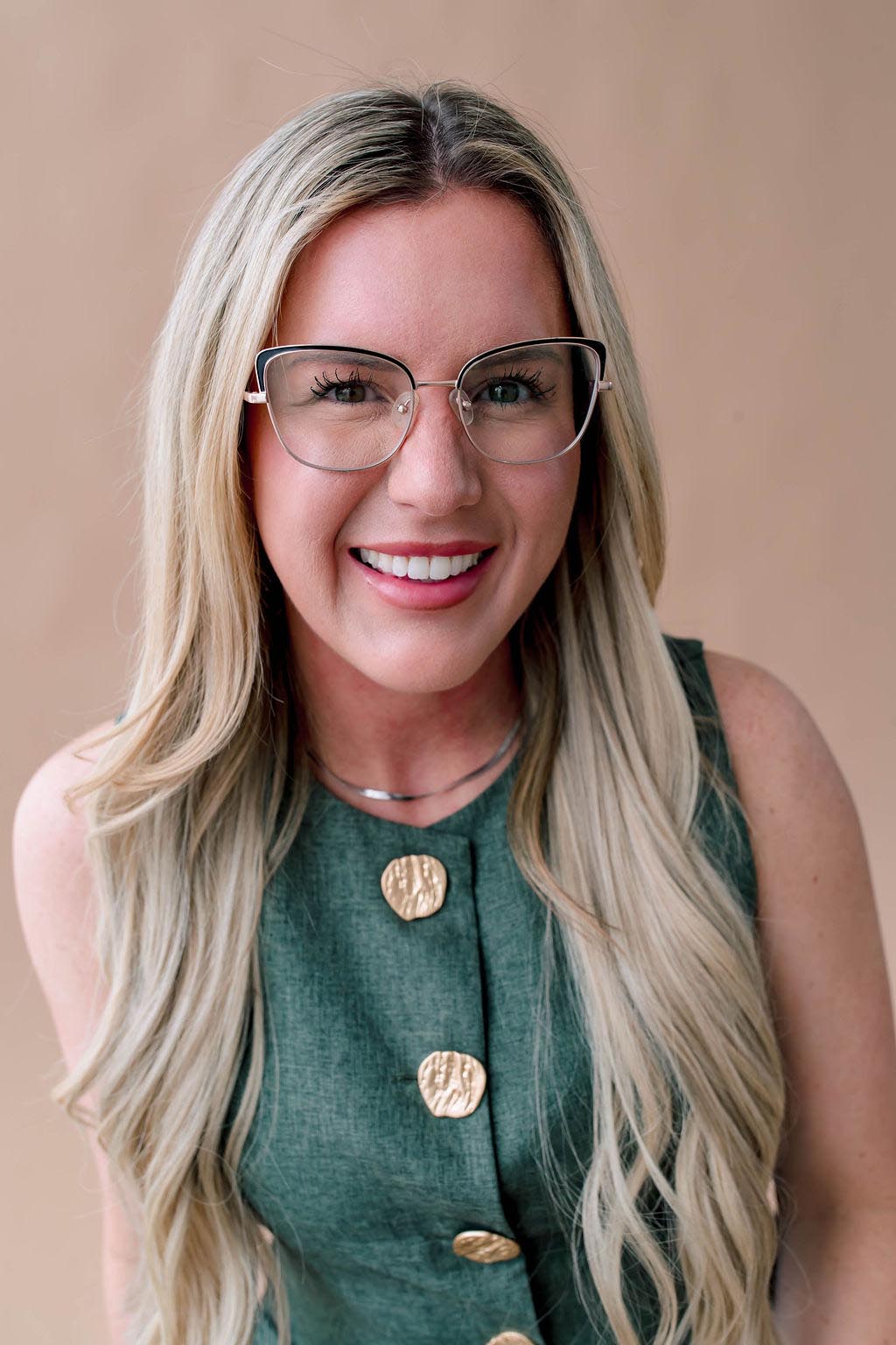 A person with glasses and long blonde hair, wearing a green top with gold buttons, smiling against a beige background.