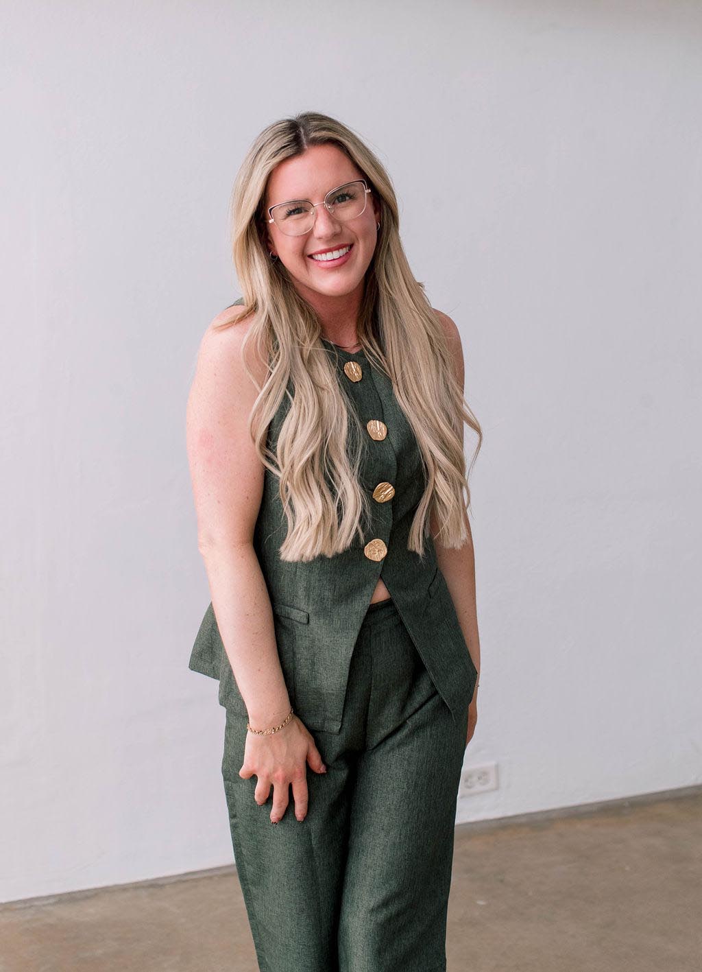 A woman with long blonde hair wearing a green suit and glasses, smiling against a light background.