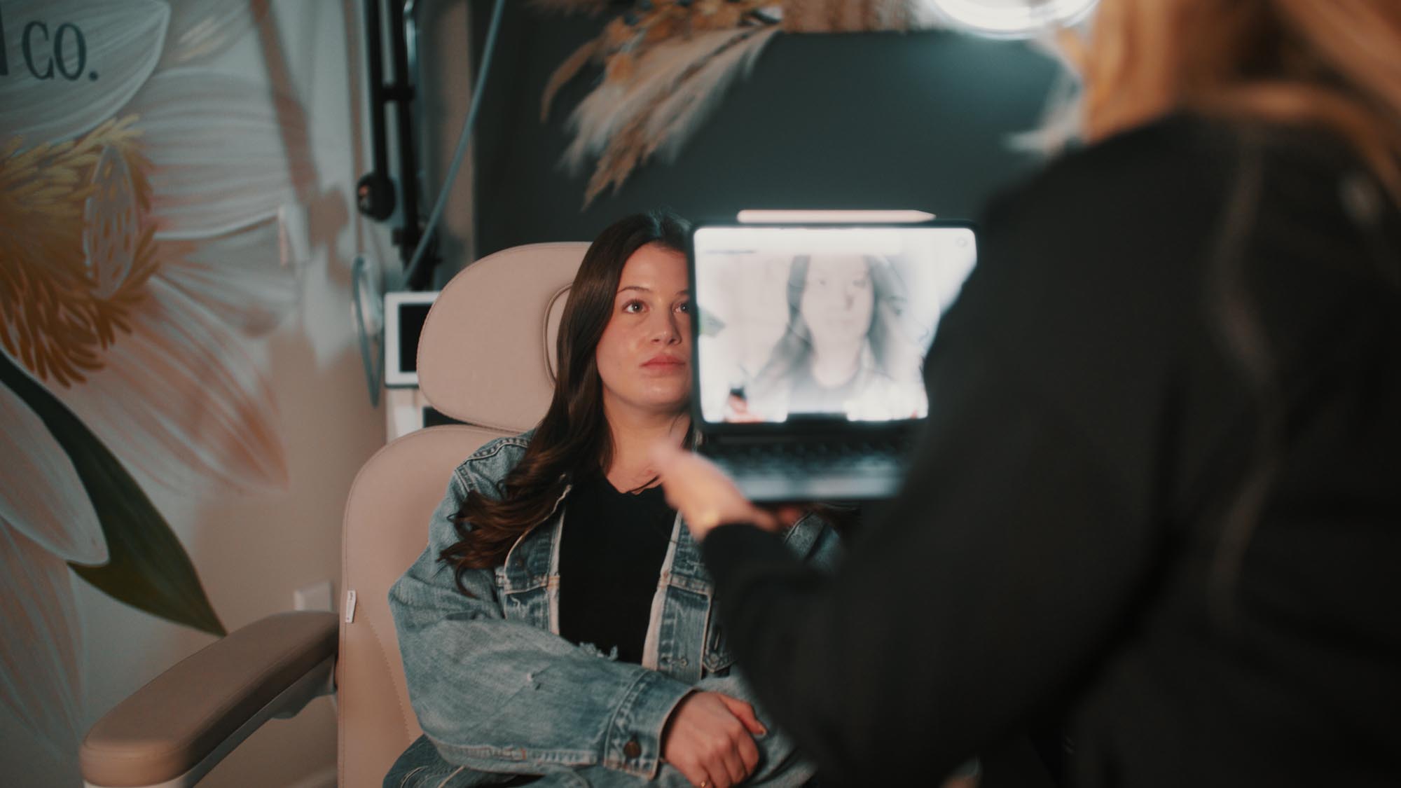 A woman in a denim jacket sitting with a laptop screen facing her in a modern interior.