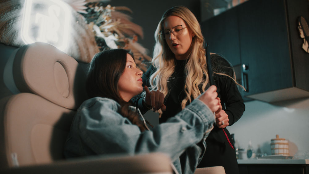 A stylist with long hair consulting a client in a salon chair.
