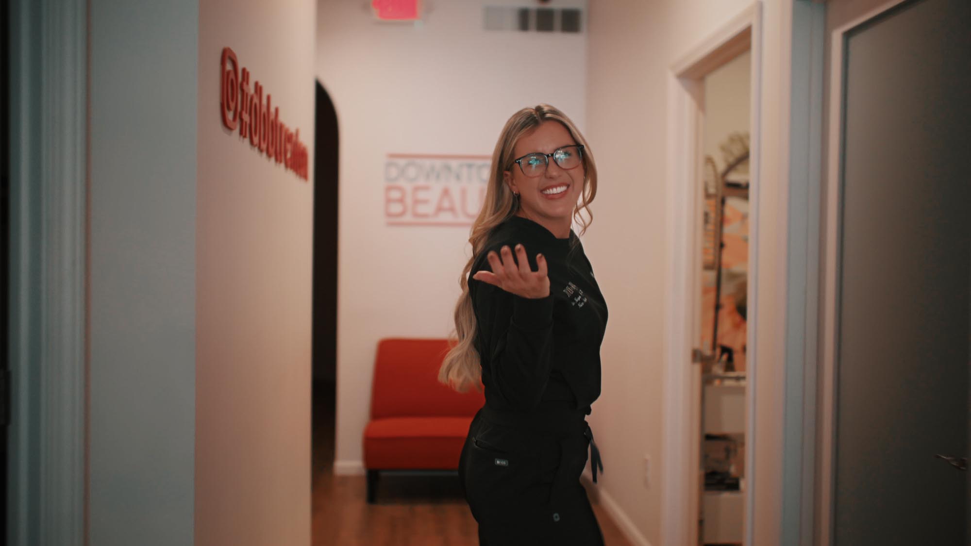 A woman with glasses smiling in a hallway, standing near a sign that reads Downtown Beauty.