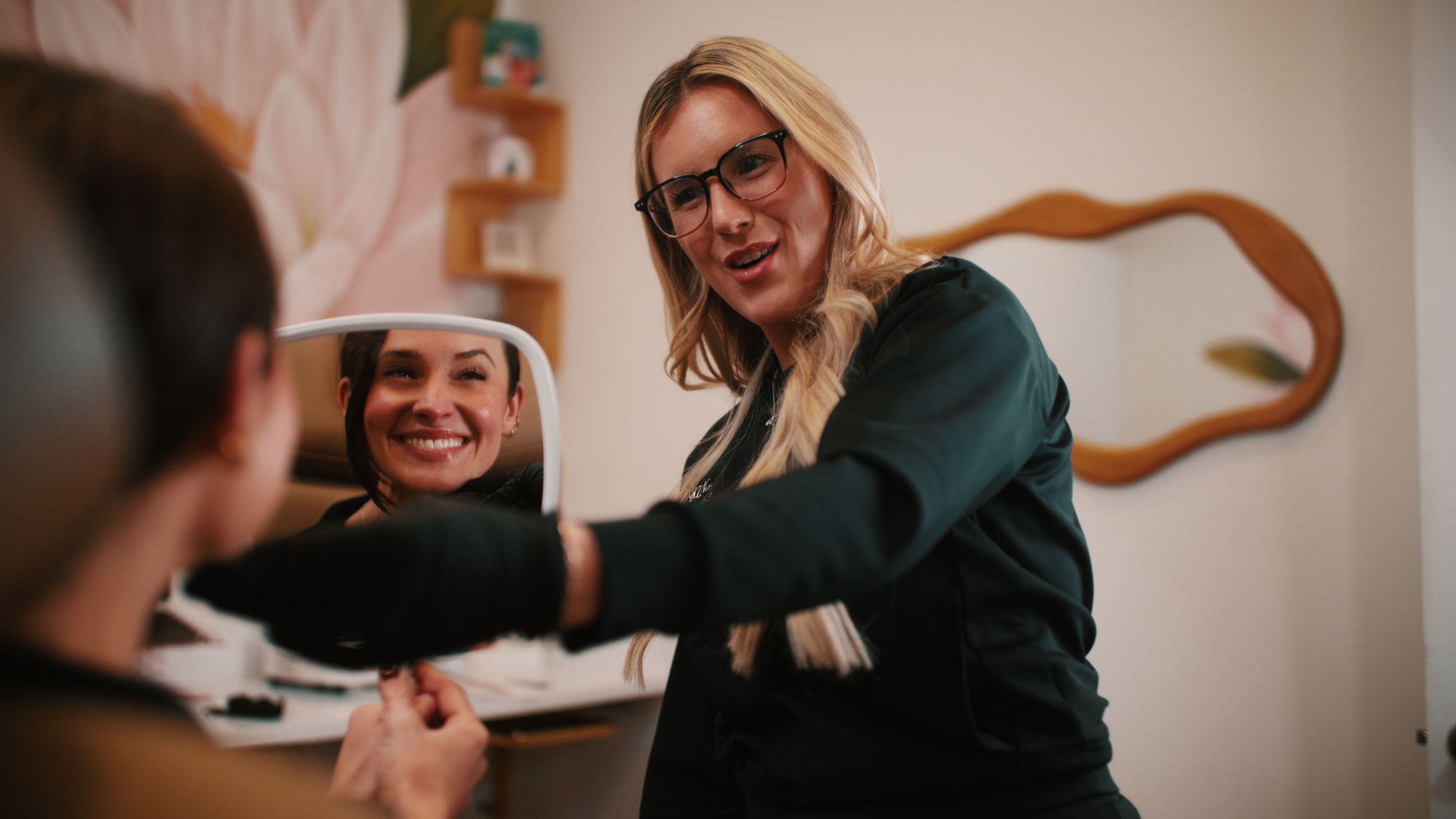 A woman with glasses and long hair shows a smiling woman her reflection in a mirror.