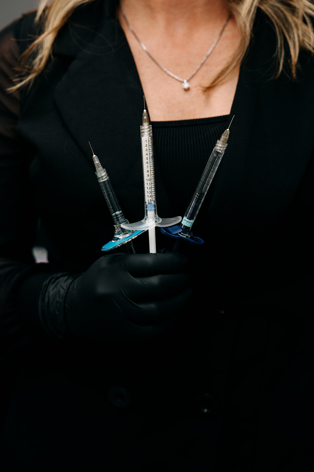 Person in black glove holding three syringes in a black outfit.