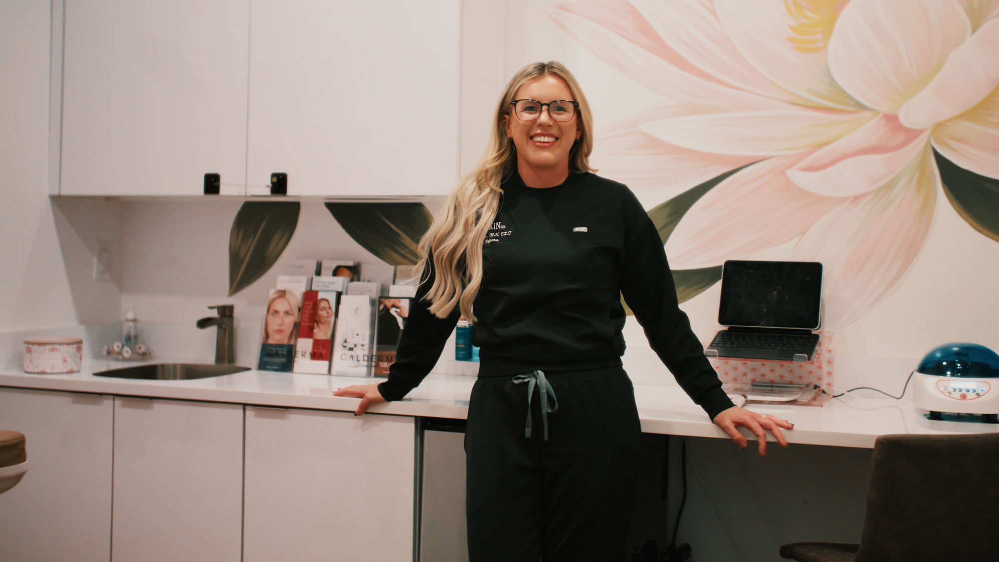 A woman smiling in a modern skincare clinic, standing by a counter with products and a laptop.