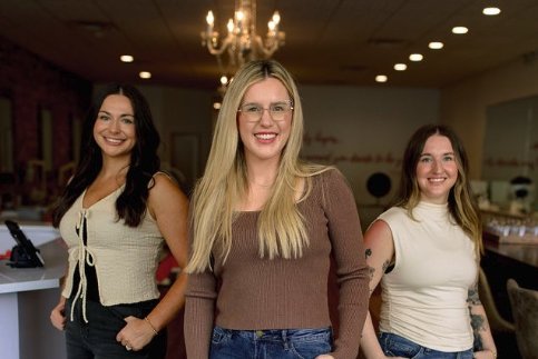Three women smiling together in a stylish indoor setting with chandeliers.